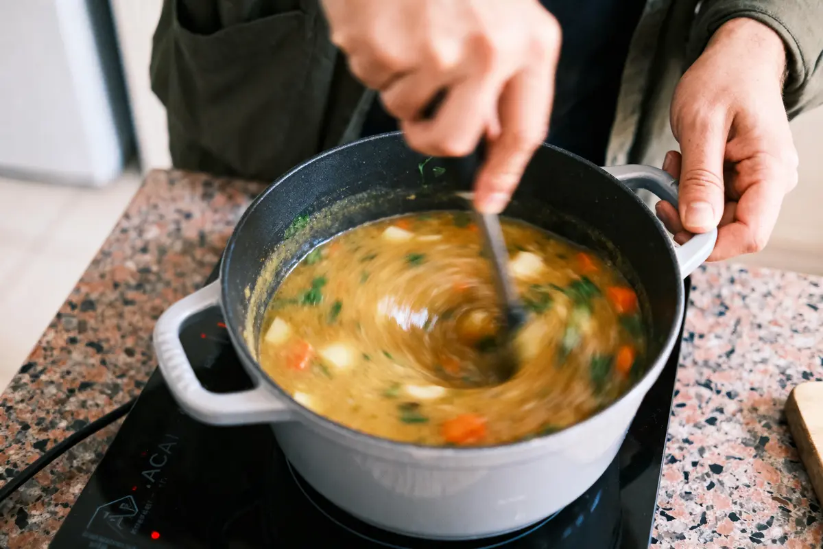 Personne mélange une soupe aux légumes dans une casserole sur plaque de cuisson, cuisine intérieure.